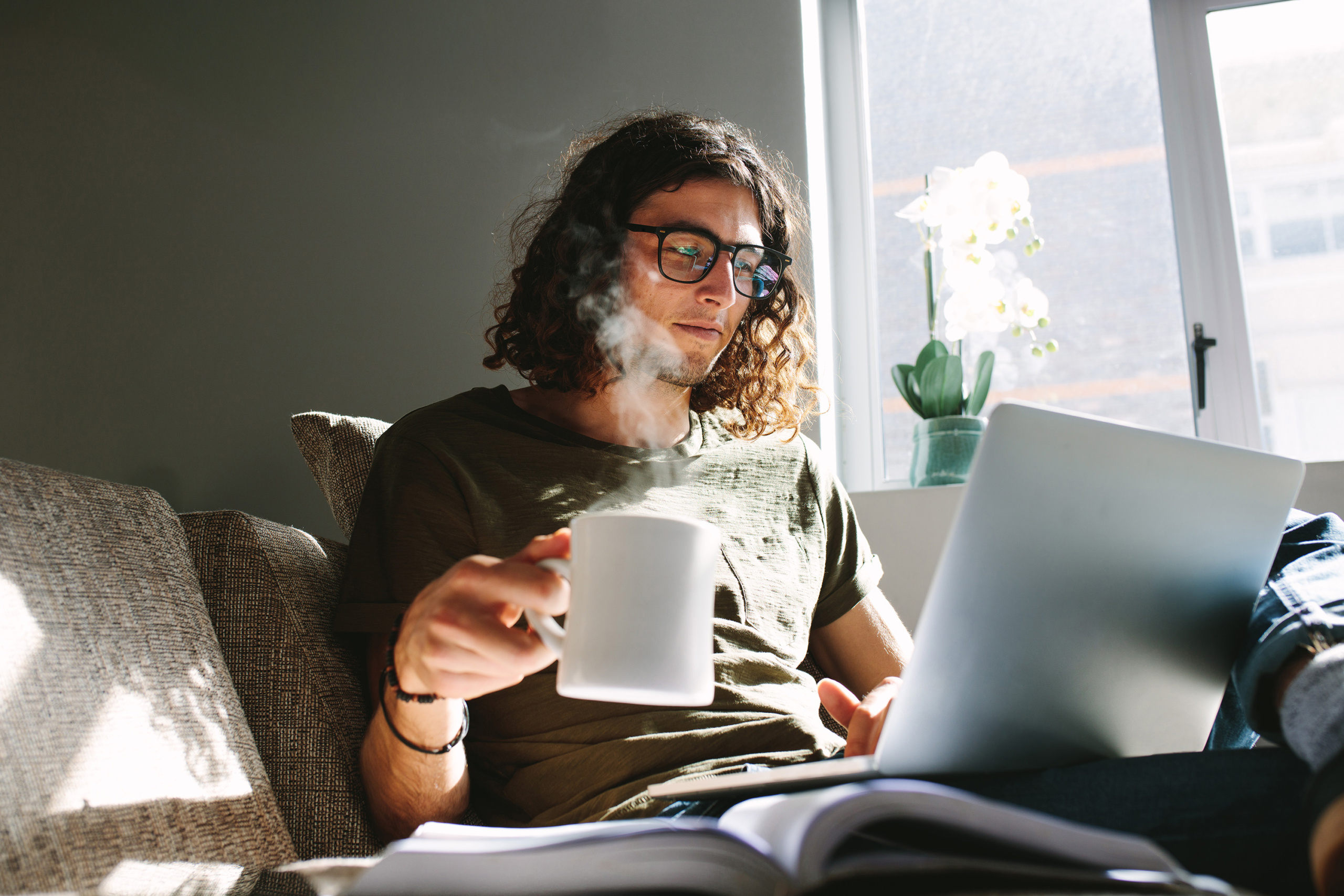 En kille sitter hemma i sin soffa med en kopp kaffe och gör en utbildningsansökning på sin laptop i knät.